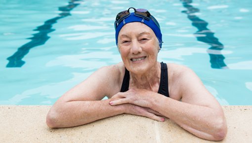 Das Foto zeigt eine ältere Frau mit Badekappe im Schwimmbad, die sich am Beckenrand abstützt